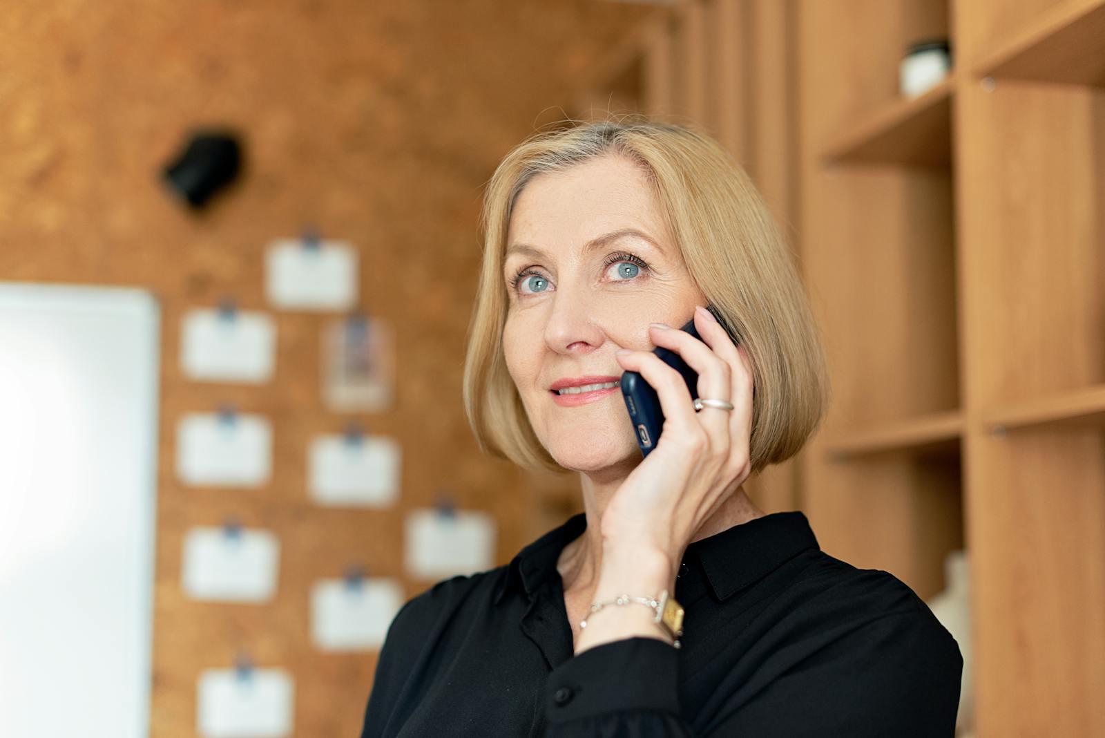 Smiling woman in an office setting, talking on a mobile phone with a confident expression.